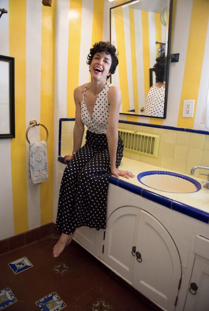 A woman in a polka dot dress sitting on a sink in a yellow bathroom.