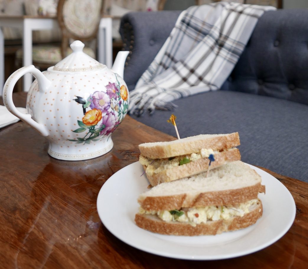 A sandwich on a plate next to a teapot.